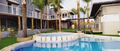 A swimming pool with a tiled edge, surrounded by palm trees and a building with balconies.