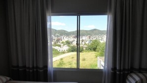Habitación de hotel con un ventanal grande, una cama y vistas a un paisaje urbano y colinas.
