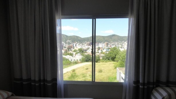 Habitación de hotel con un ventanal grande, una cama y vistas a un paisaje urbano y colinas.