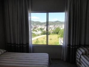 A hotel room with a large window, a bed, and a view of a cityscape and hills.
