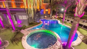 Una piscina con un jacuzzi, rodeada de palmeras y un edificio con ventanas iluminadas.