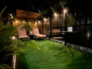 Eine Außenpatio-Terrasse mit Kunstgrashimmel, zwei Lounge-Stühlen, einem kleinen Tisch und einem Blumentopf.