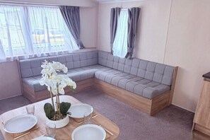 A room with a grey sofa, a dining table set with plates and glasses, and a potted plant. - 2-bed Caravan - Sleeps 6 - Pet Friendly - Parking (Skegness)