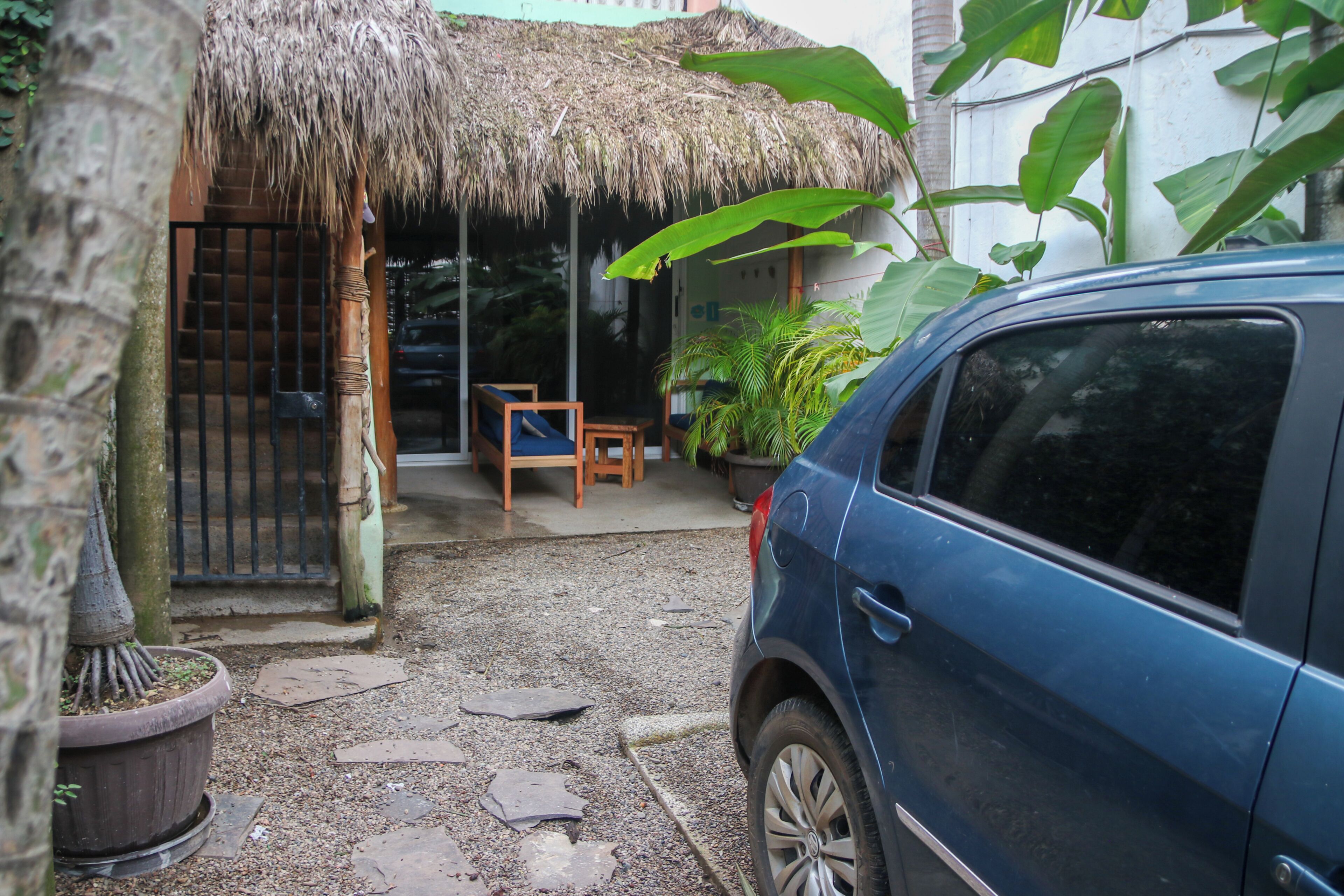 A blue car parked in a driveway with a thatched-roof structure, a wooden bench, and potted plants.