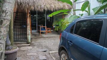 A blue car parked in a driveway with a thatched-roof structure, a wooden bench, and potted plants.