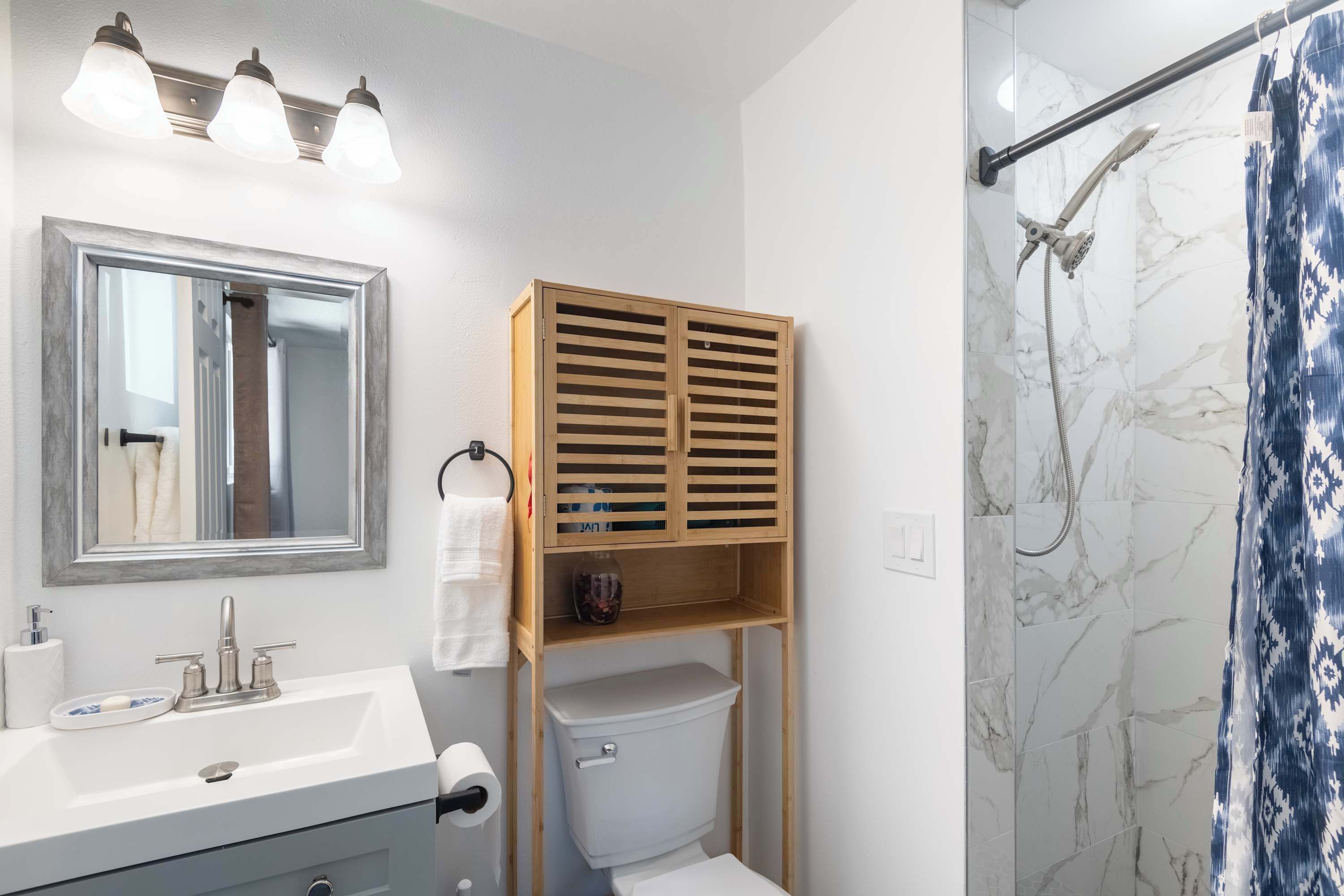 Un baño con lavamanos blanco, espejo, mueble de madera, inodoro, ducha y azulejos de mármol.