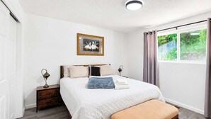 A bedroom with a bed, a bedside table, a lamp, a picture frame, a window with curtains, and a view of greenery outside. - Seattle Hillside Escape with Decks & Scenic Views (Seattle)