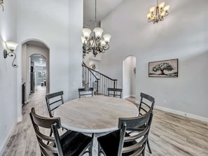 A dining area with a round table, six chairs, a chandelier, and a staircase leading to another level.