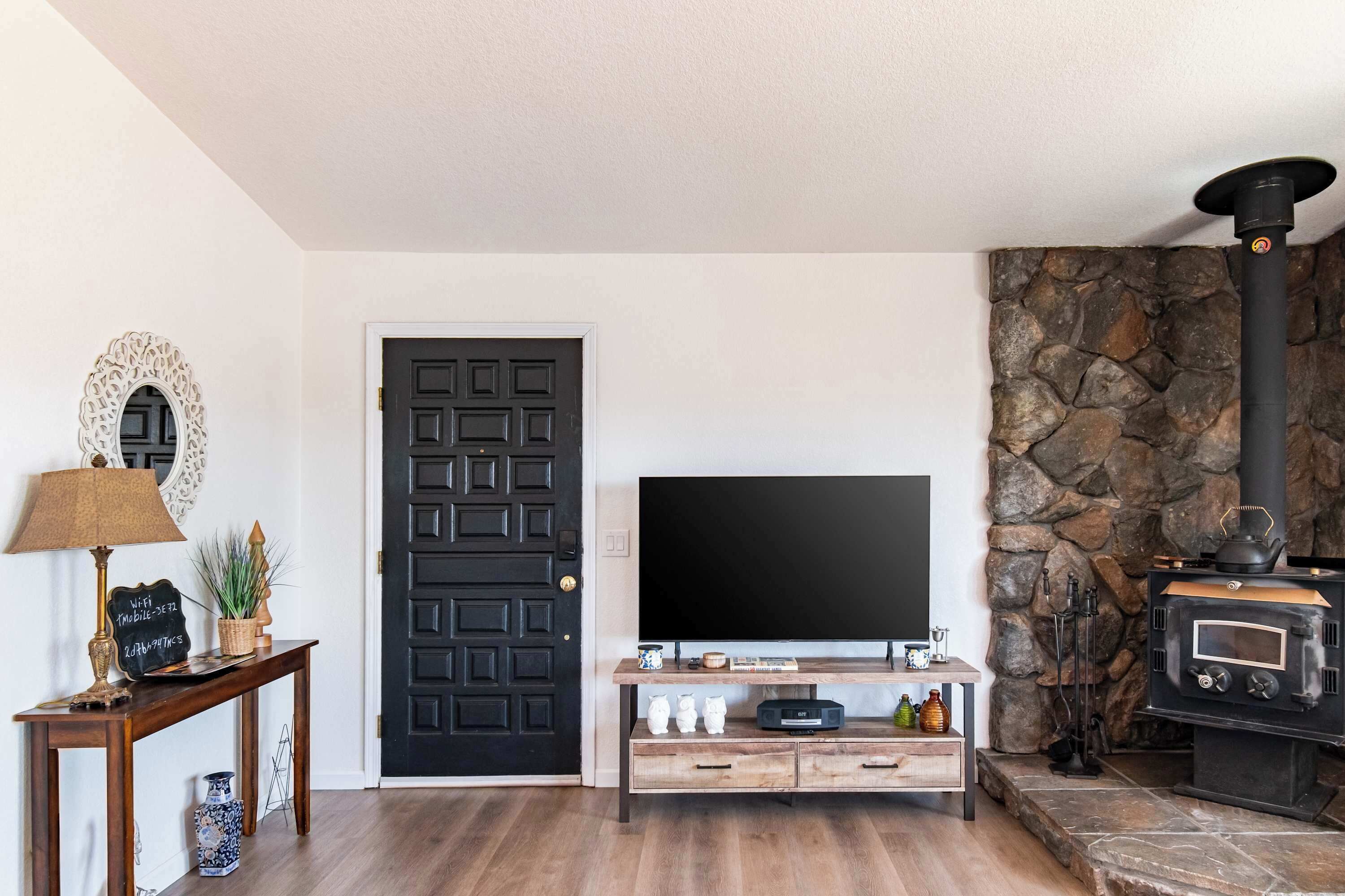 A living room with a black door, a flat-screen TV, a wooden console table, and a stone fireplace.