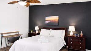 Un lit bien fait, avec du linge de lit blanc, une tête de lit en bois, deux tables de chevet avec des lampes, un bureau et un ventilateur de plafond.
