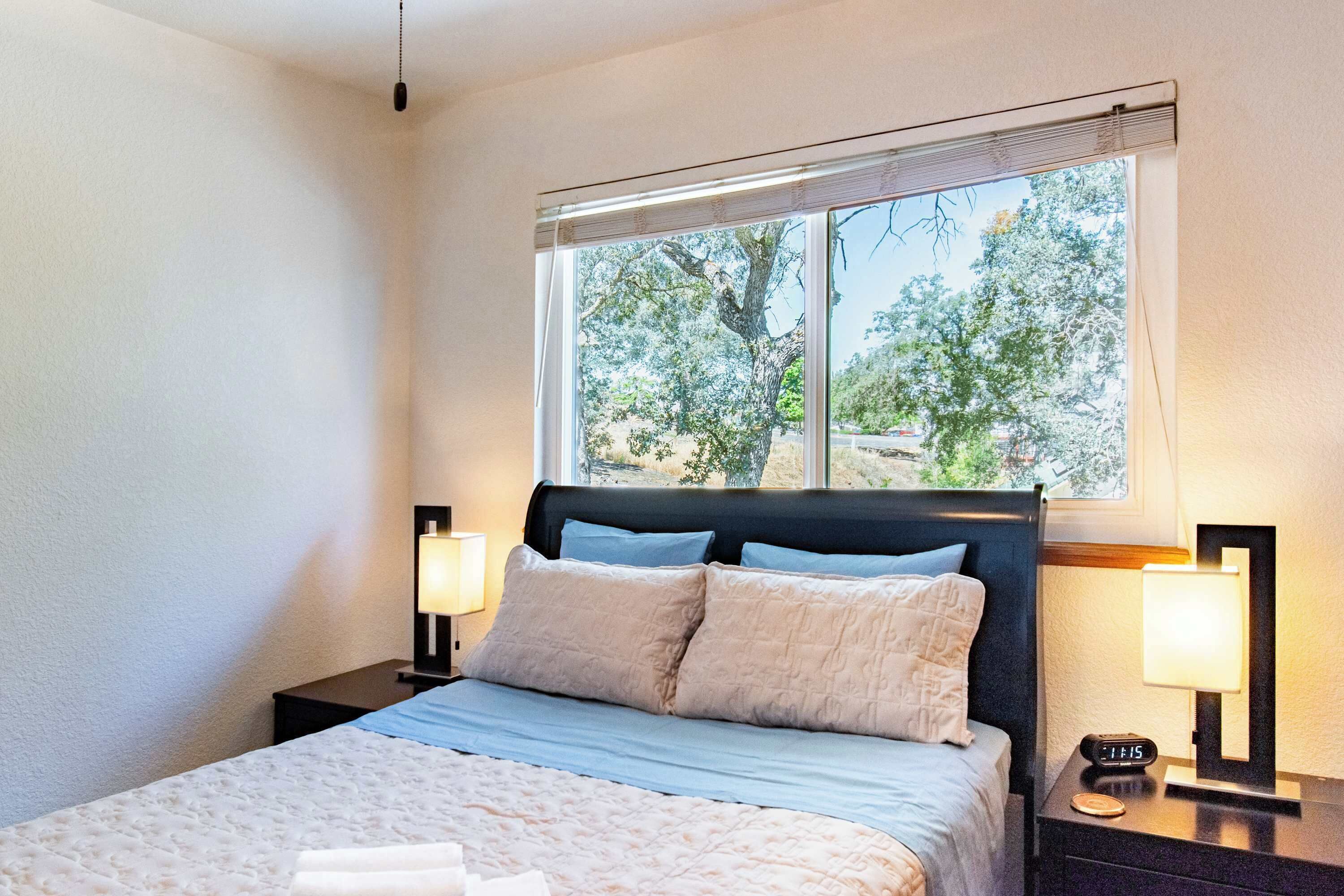 A bedroom with a bed, two bedside tables, a window with a view of trees, and a nightstand with a lamp.