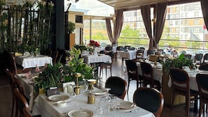En restaurang med bord som är dukade för middag, utsikt över byggnader utanför och ett tak med synliga ventilationskanaler.