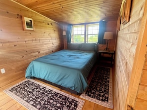 A wooden cabin room with a bed, a window, a nightstand, and a lamp.