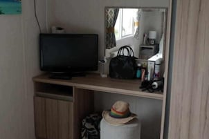 A room with a television on a wooden shelf, a mirror, a small table with a black bag, and a hat on a white cylindrical object.