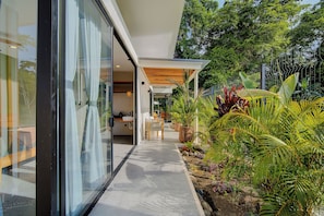 Una casa moderna con fachada de vidrio, una pérgola de madera y un jardín con plantas tropicales.