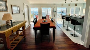 A dining area with a wooden table, chairs, and a bench. There is a kitchen area with a bar counter and stools. The room has large windows with a view of the sea. - Big Waterfront House! Fishing Dock, Arcade, Garage Parking, Walk to Beach! (Norfolk)