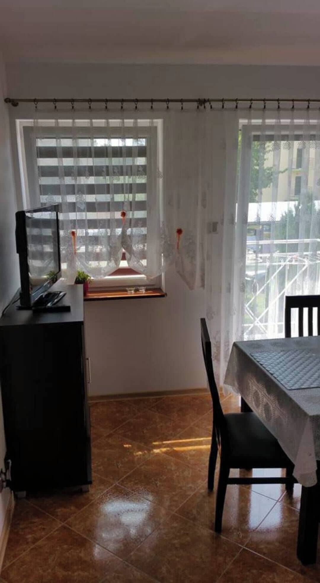 Une pièce comprenant une table à manger, des chaises, un téléviseur posé sur un meuble et une fenêtre avec des stores.