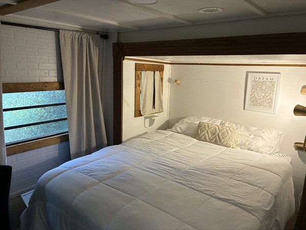 Ein Schlafzimmer mit einem Bett, einem fensterbelaubten Fenster, einem Holzkopfende, einer Wandlampe und einem gerahmten Zitat an der Wand.