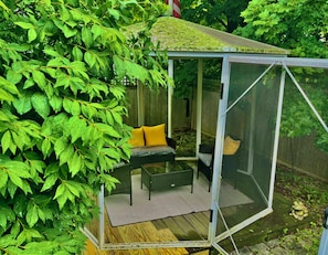 A covered outdoor seating area with a table and chairs, surrounded by lush greenery.