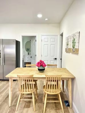 A dining area with a wooden table, four chairs, a vase with flowers, a refrigerator, and a door with a mirror.