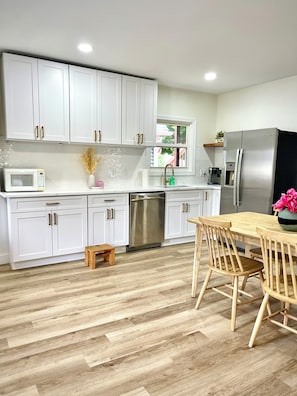 A modern kitchen with white cabinets, a stainless steel refrigerator, a wooden dining table with chairs, and a small wooden stool.