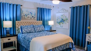 Quarto com cabeceira estofada, cama com roupa de cama azul e branca, duas mesas de cabeceira com abajures e cortinas azuis.