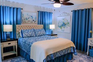 A bedroom with a tufted headboard, a bed with blue and white bedding, two bedside tables with lamps, and blue curtains. - Nanny`s Nest - A Gentle Place to Land (Nederland)