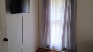 A room with a flat-screen TV mounted on the wall, a door with a visible handle, and a window with sheer curtains.