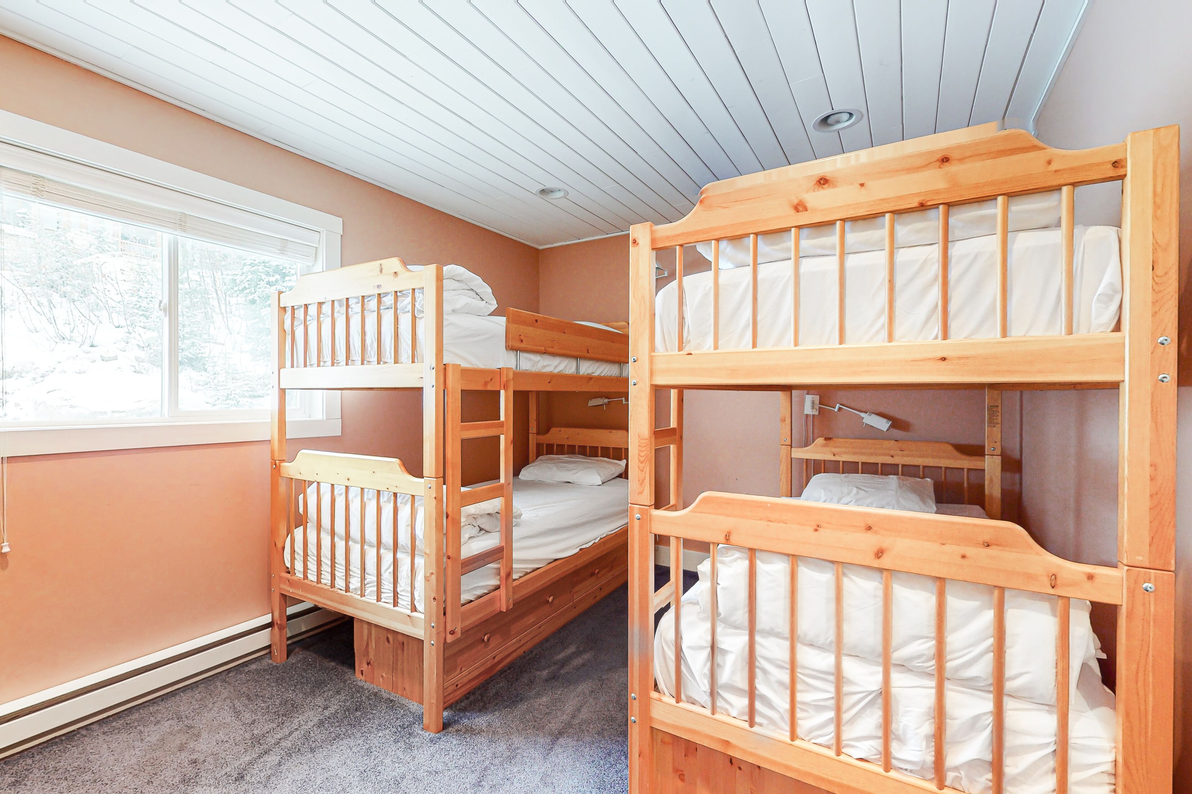 A room with three wooden bunk beds, a window with blinds, and a ceiling with wooden panels.