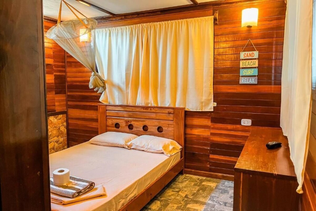 A wooden room with a bed, a hanging mosquito net, a wall-mounted light, and a sign with words 'SAND BEACH SURF RELAX'.