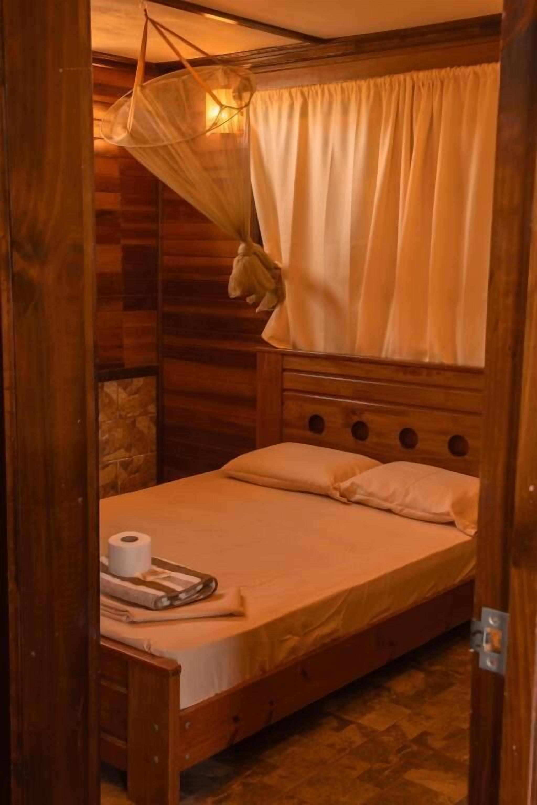 Ein Zimmer mit Holzboden, einem Bett, einem hängenden Moskitonetz und einer Toilettenpapierrolle auf dem Bett.