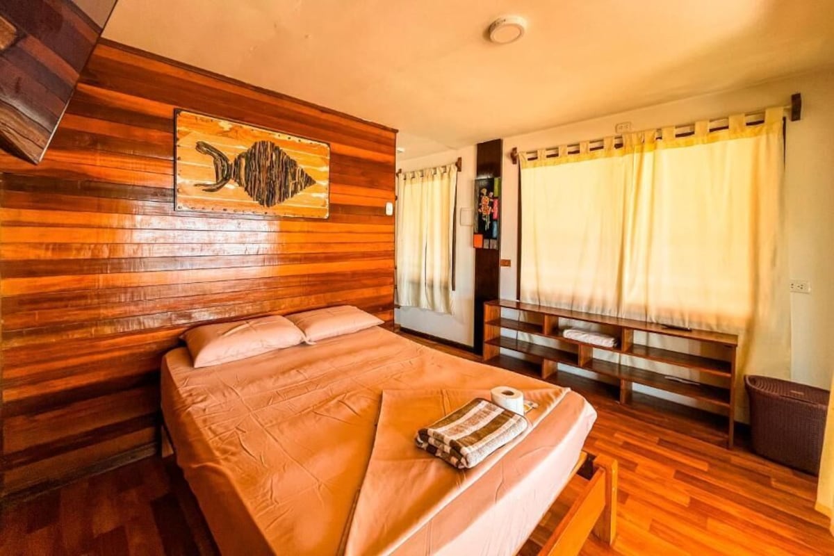 A room with a wooden headboard, a bed with a brown bedspread, a painting on the wall, and curtains on the windows.