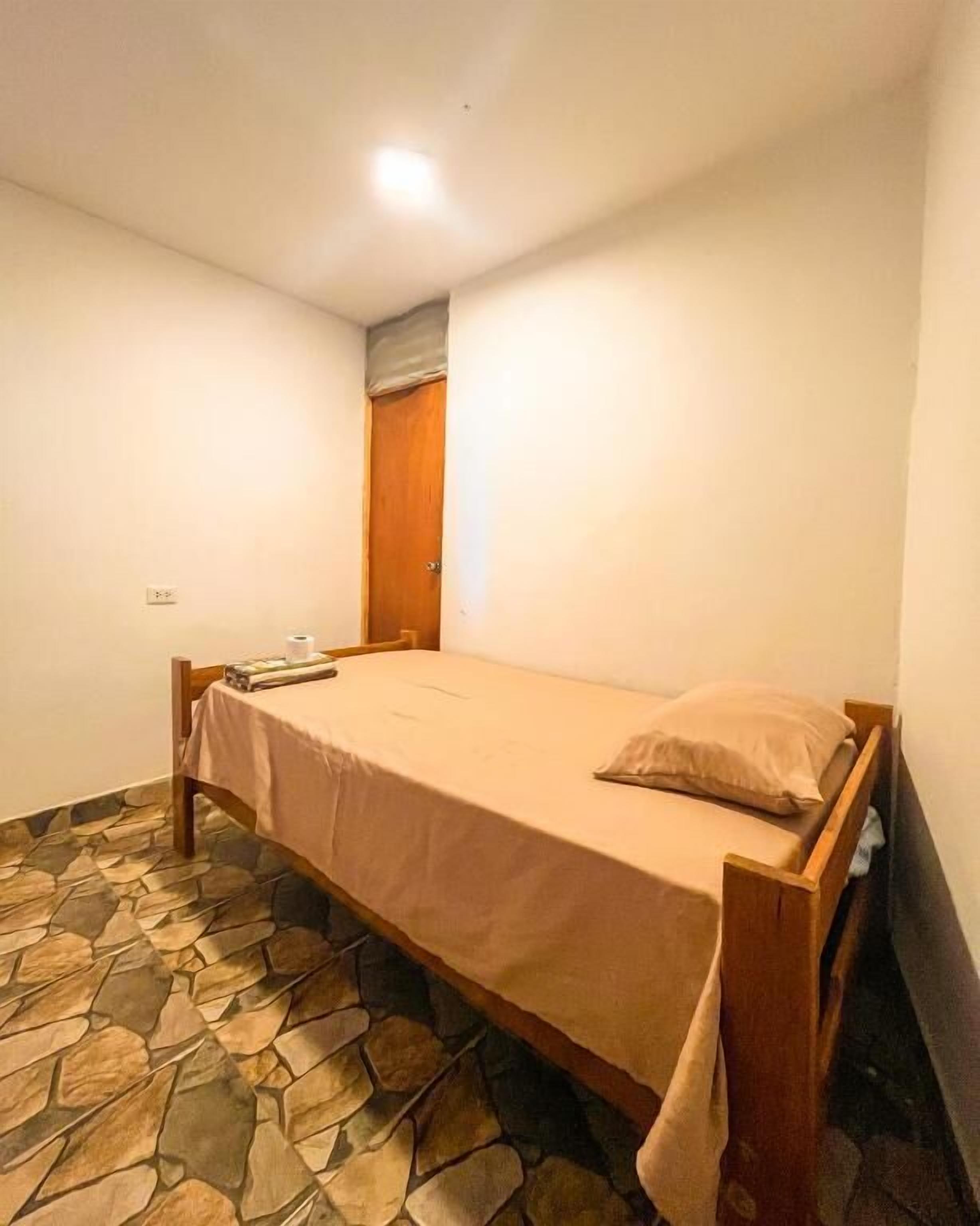 A single bed with a wooden frame, a stone-patterned floor, and a plain wall.