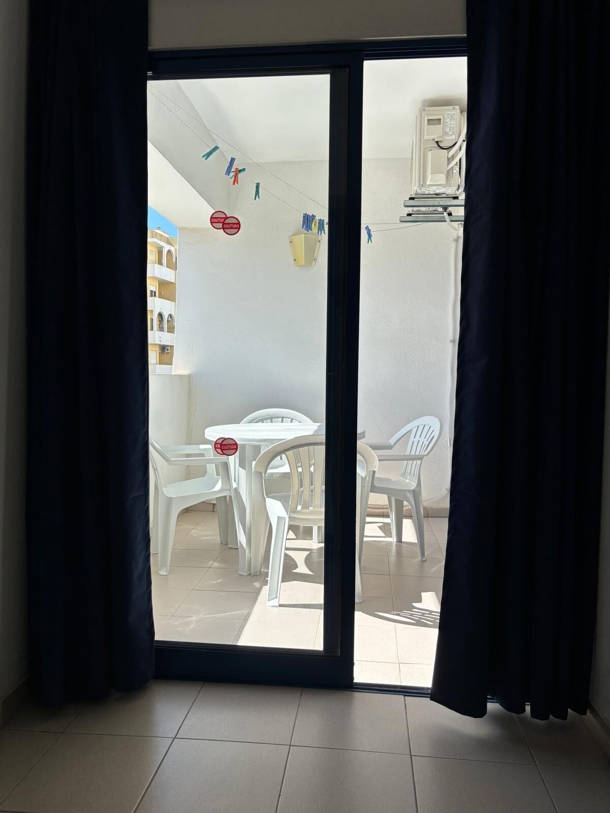 Une pièce dotée d’une porte-fenêtre coulissante donnant sur un espace extérieur avec une table et des chaises blanches, un petit meuble blanc et un appareil fixé au plafond.