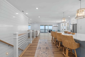 A modern kitchen with white cabinetry, a wooden floor, and a dining area with wicker chairs. - Sea Dreams- Ocean Isle Beach (Ocean Isle Beach)