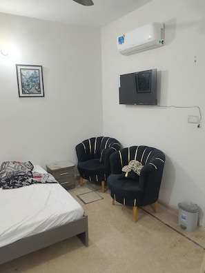 A bedroom with a bed, two armchairs, a TV, and an air conditioner.