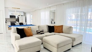 A modern living room with a sectional sofa, ottomans, a pool view, and a kitchen in the background. - Designer house near Disney with private pool, water views, and custom pool table (Kissimmee)