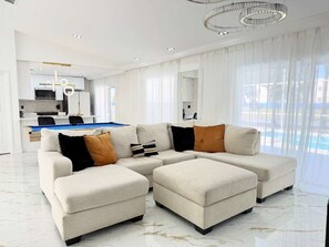 A modern living room with a sectional sofa, ottomans, a pool view, and a kitchen in the background. - Designer house near Disney with private pool, water views, and custom pool table (Kissimmee)