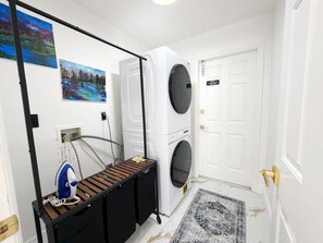 Ein Waschraum mit zwei übereinandergestellten Waschmaschinen, einem Wäscheständer und einem an der Wand befestigten Bügelbrett.