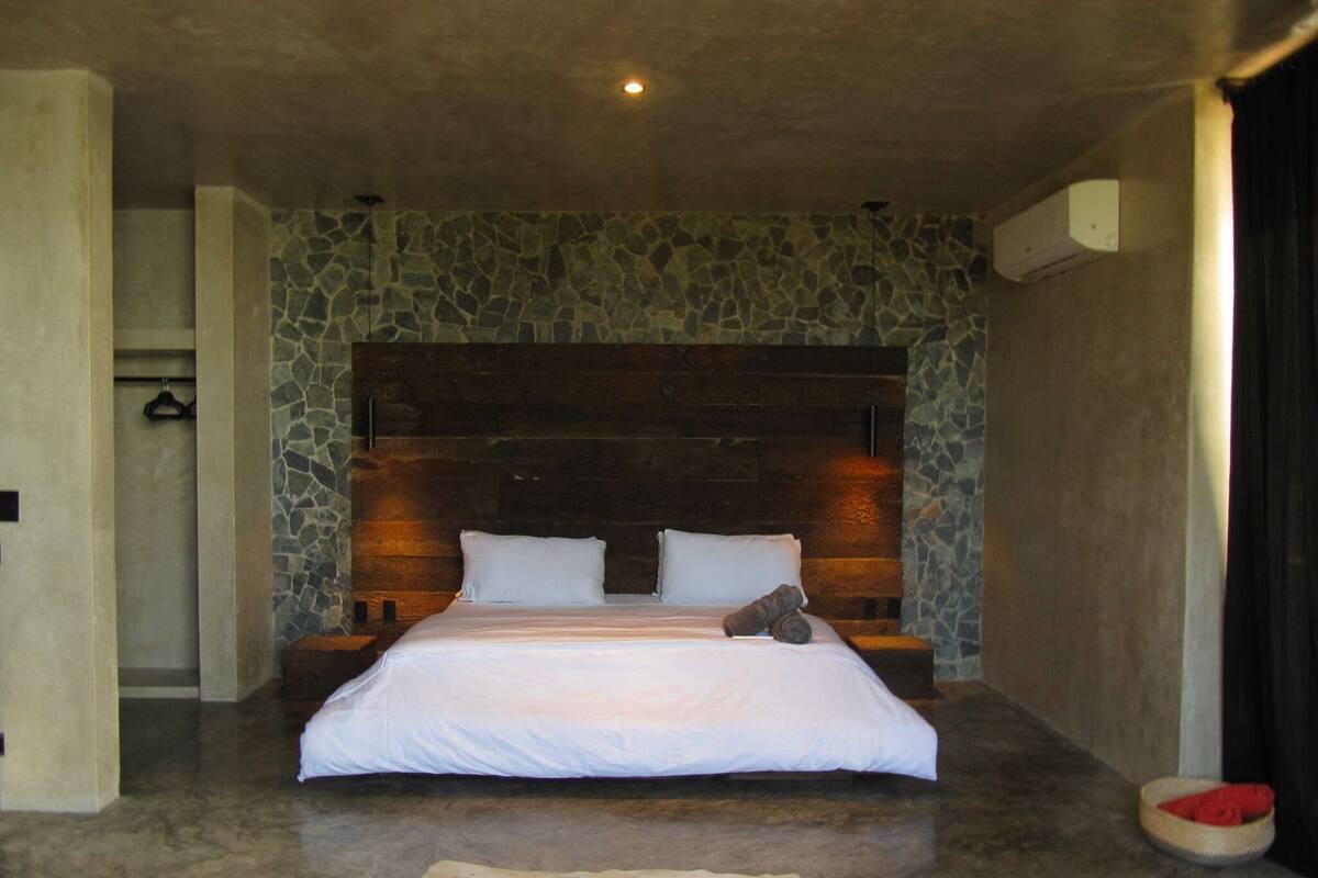 Un dormitorio con pared de piedra, cabecera de madera, cama con ropa de cama blanca y una alfombra pequeña.