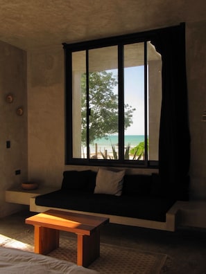 A room with a large window, a sofa, a wooden table, and a view of the beach. - Hotel Amadeo (Isla Holbox)