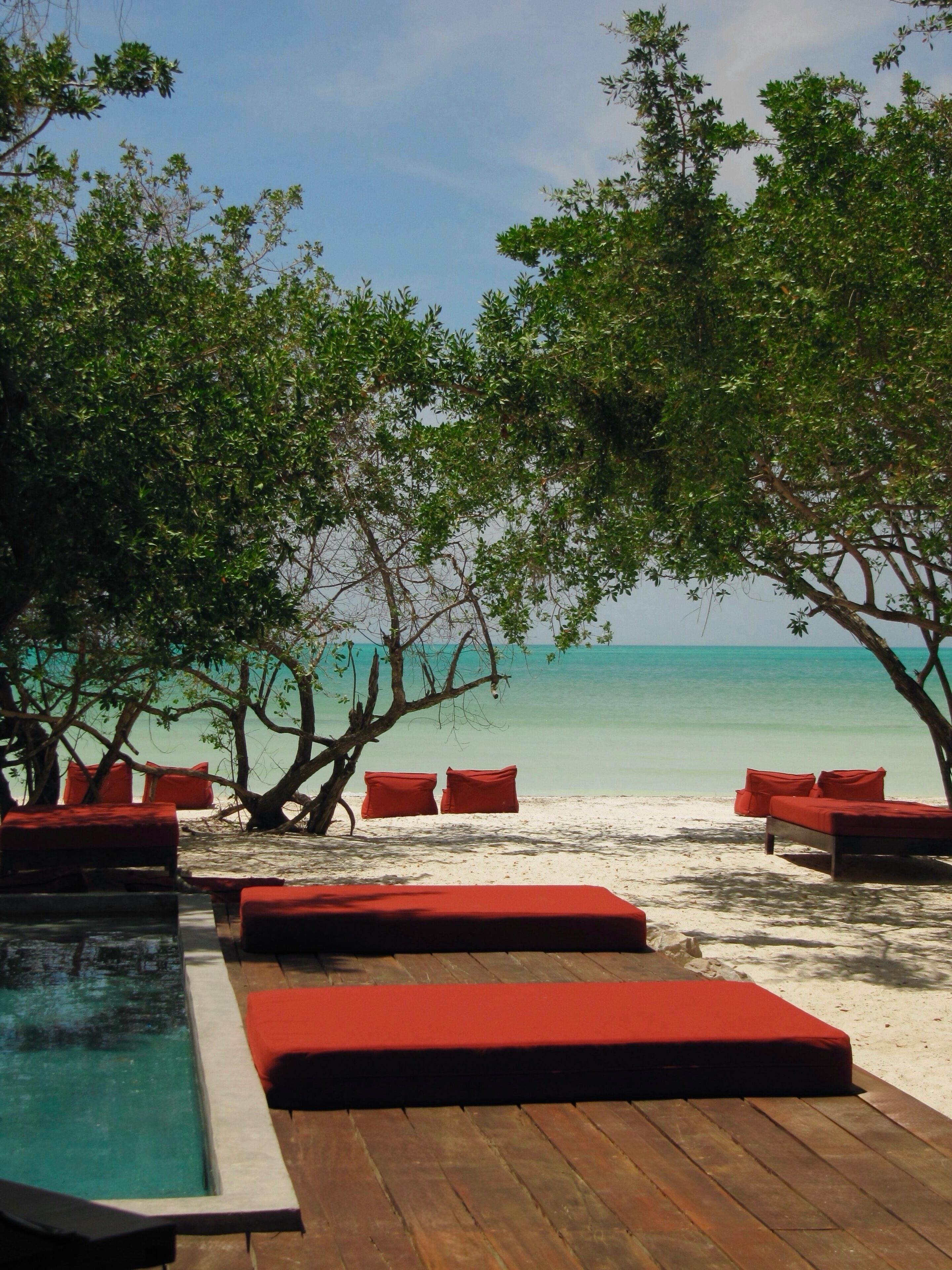 A beachside lounge area with wooden flooring, red cushioned seating, and a pool.