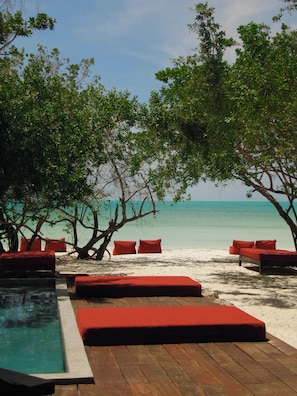 Un área de descanso frente a la playa con piso de madera, asientos acolchados rojos y una piscina.