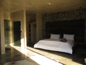 Un dormitorio con pared de piedra, cama con ropa blanca, cabecera de madera y estantería empotrada.