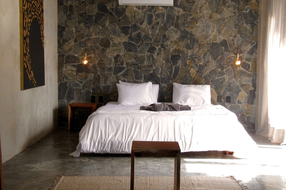 Un dormitorio con pared de piedra, una cama con ropa de cama blanca, dos mesitas de noche y una lámpara colgante.