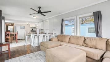 Una sala de estar con un sofá seccional beige, una mesa de comedor con sillas, un ventilador de techo y vistas a un edificio azul en el exterior.