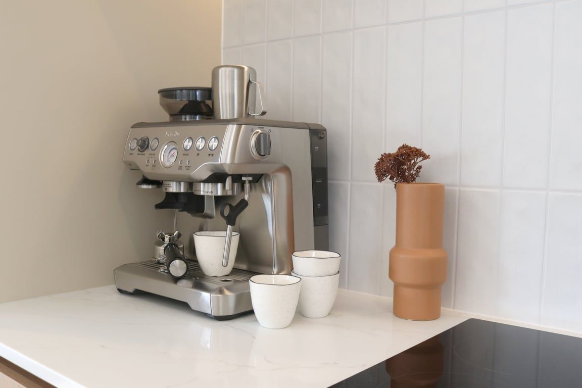 Una cocina con una cafetera, tres tazas blancas y un jarrón marrón con flores secas.