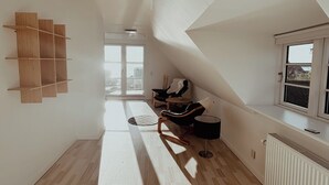 Une pièce spacieuse avec un plafond en pente, un radiateur, une fenêtre et un coin salon comprenant une chaise et une table.