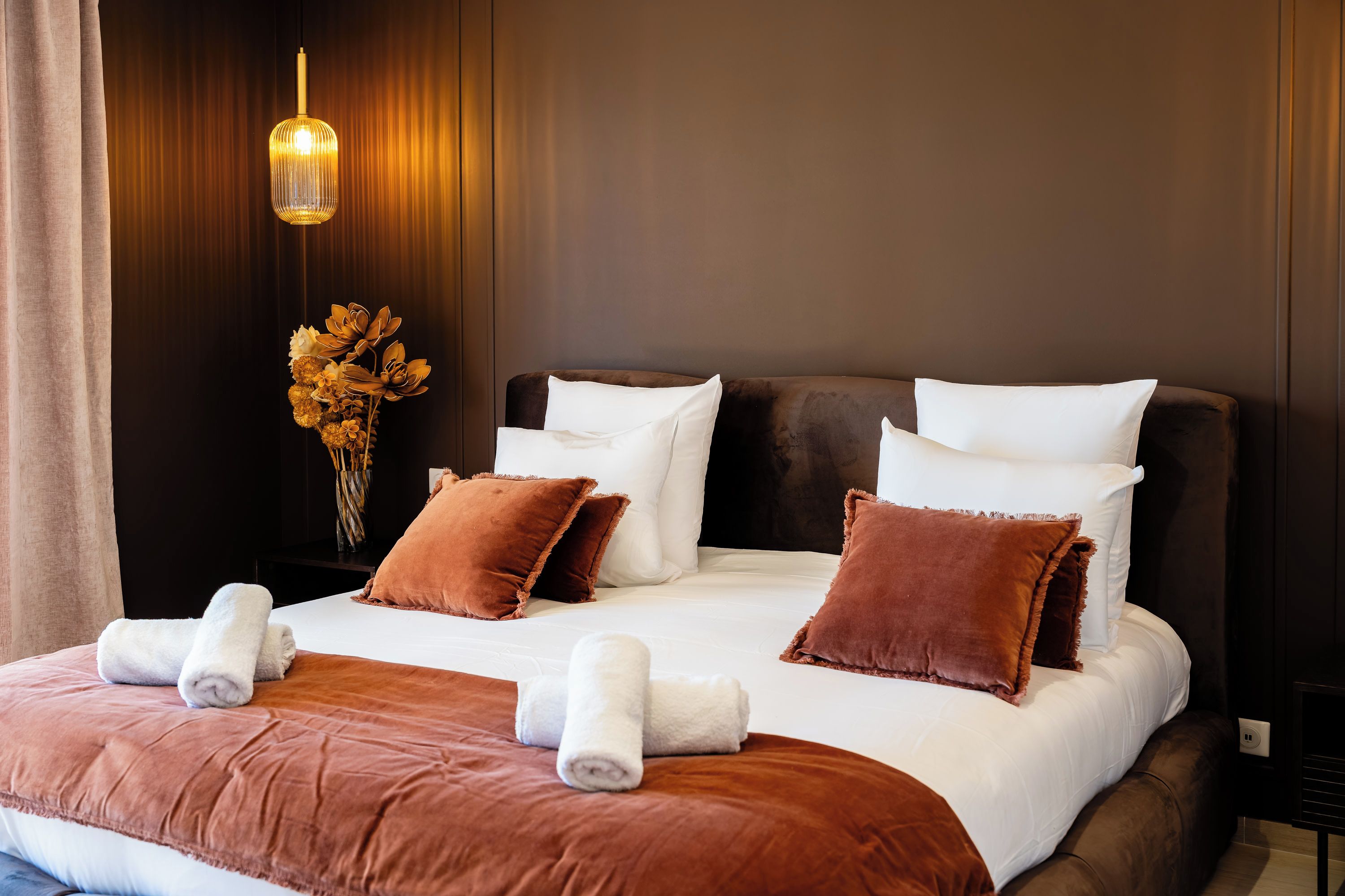 A neatly made bed with brown and white bedding, a vase with dried flowers, and a hanging light fixture.
