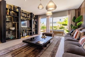 A modern living room with a sofa, coffee table, bookshelf, and large windows. - Villa Maya · Rooftop beach access heated pool (Agde)